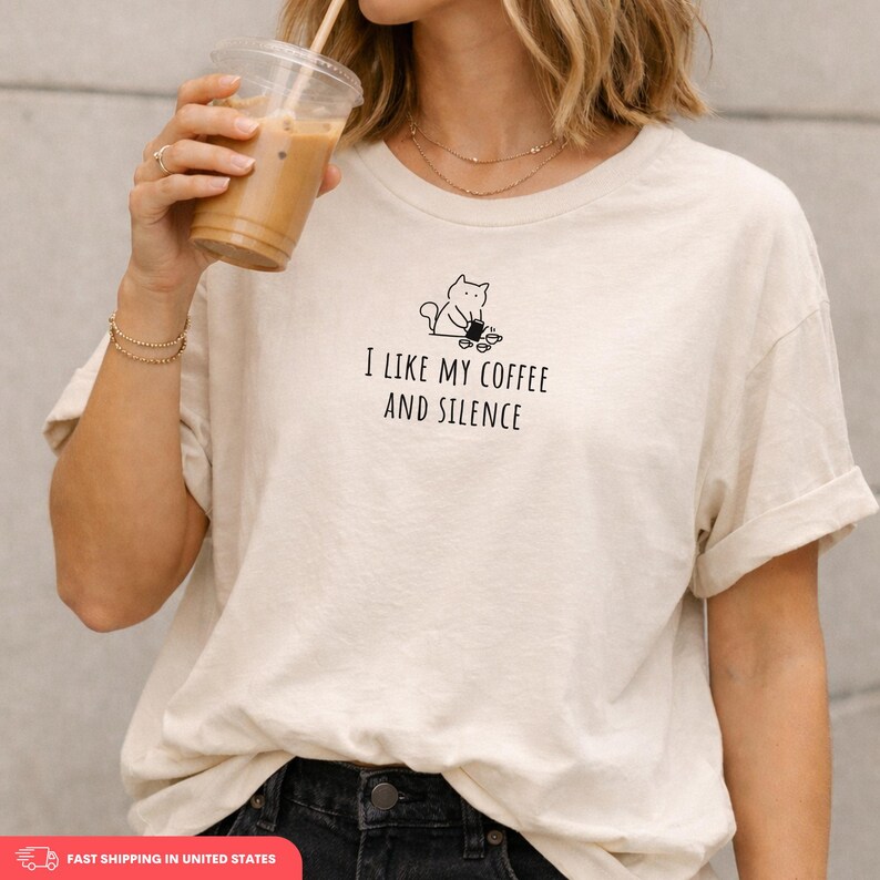 May include: Cream-colored t-shirt with the text "I LIKE MY COFFEE AND SILENCE" in black. The shirt features a small black graphic of a cat with coffee cups. The sleeves are rolled up.