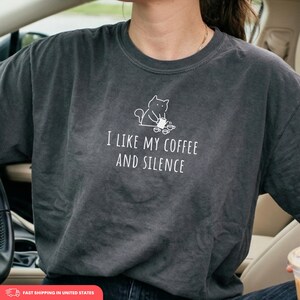 May include: Dark gray t-shirt with the text "I LIKE MY COFFEE AND SILENCE" in white. Above the text is a white illustration of a cat holding a coffee cup. The shirt is a casual wear item.