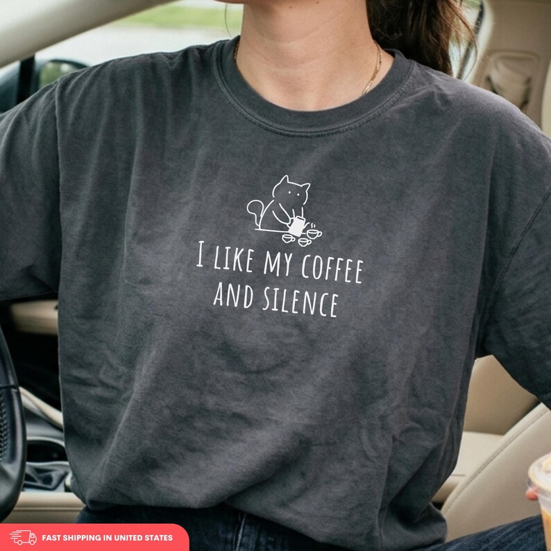 May include: Dark gray t-shirt with the text "I LIKE MY COFFEE AND SILENCE" in white. Above the text is a white illustration of a cat holding a coffee cup. The shirt is a casual wear item.