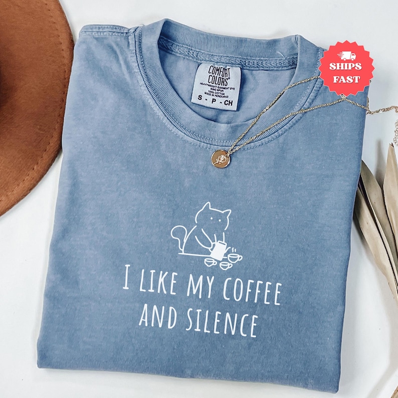 May include: A blue Comfort Colors t-shirt with the phrase "I LIKE MY COFFEE AND SILENCE" in white. A gold necklace with a pendant rests on the shirt. A white cat graphic is above the text. The shirt is size small.