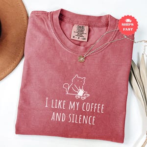 May include: A dusty rose Comfort Colors t-shirt with the phrase "I LIKE MY COFFEE AND SILENCE" and a cat graphic. A silver necklace with a pendant rests on the shirt. The shirt is folded, with a brown hat visible in the background.