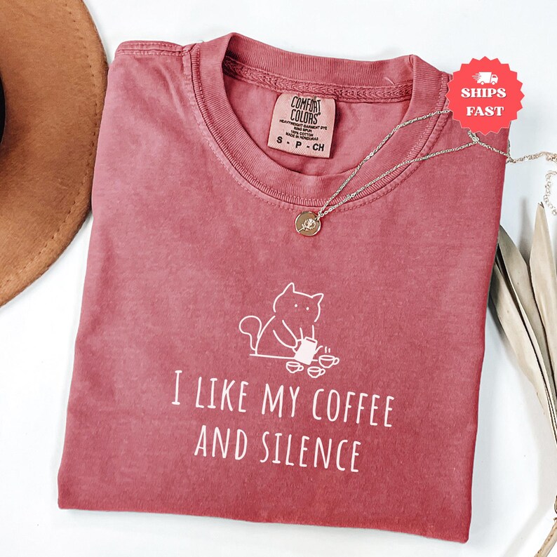 May include: A dusty rose Comfort Colors t-shirt with the phrase "I LIKE MY COFFEE AND SILENCE" and a cat graphic. A silver necklace with a pendant rests on the shirt. The shirt is folded, with a brown hat visible in the background.