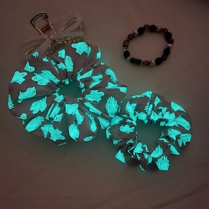 Op de afbeelding: Twee grijze scrunchies met een gloeiend teal spookpatroon, een kralenarmband en een doorzichtige sleutelhanger met een strik. De scrunchies zijn in verschillende maten. De armband heeft bruine, zwarte en zilveren kralen.
