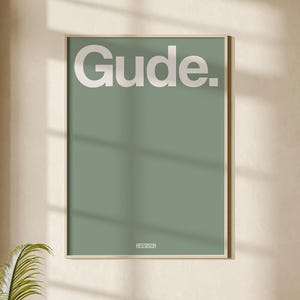 Könnte beinhalten: Ein gerahmter Kunstdruck mit dem Wort "Gude" in weißen Buchstaben auf einem salbeigrünen Hintergrund hängt über einem beigen Sofa. Zwei runde Couchtische mit Holzsockeln und Marmorplatten stehen vor dem Sofa. Eine Stehlampe und eine Topfpflanze befinden sich ebenfalls im Raum.