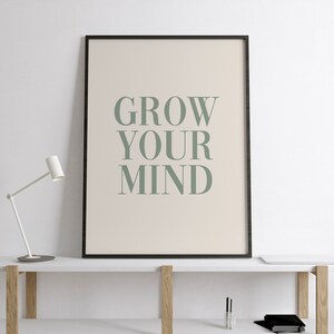 May include: A black framed print with the text "GROW YOUR MIND" in green on a beige background. The print is hanging on a white wall above a white shelf with a lamp and other items.