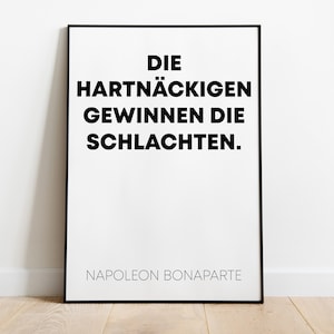May include: A framed print with a white background and black text. The text reads "DIE HARTNÄCKIGEN GEWINNEN DIE SCHLACHTEN." and "NAPOLEON BONAPARTE" at the bottom. The frame is black.