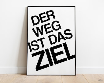 May include: Black framed art print with the German phrase "DER WEG IST DAS ZIEL" in bold, black letters on a white background. The text is arranged diagonally. The artwork is displayed on a light wood floor against a white wall.