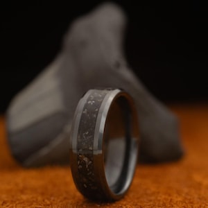 May include: A black ring with a dark, textured center band and a silver-colored border. The ring is set against a blurred, dark background and rests on an orange surface. The ring's design suggests a focus on jewelry and accessories.