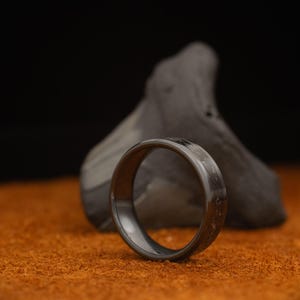 Peut inclure: Une bague grise fonc&eacute;e avec une surface textur&eacute;e, sur un fond flou. La bague est faite d'un mat&eacute;riau sombre et repose sur une surface orange. La bague est un bijou.