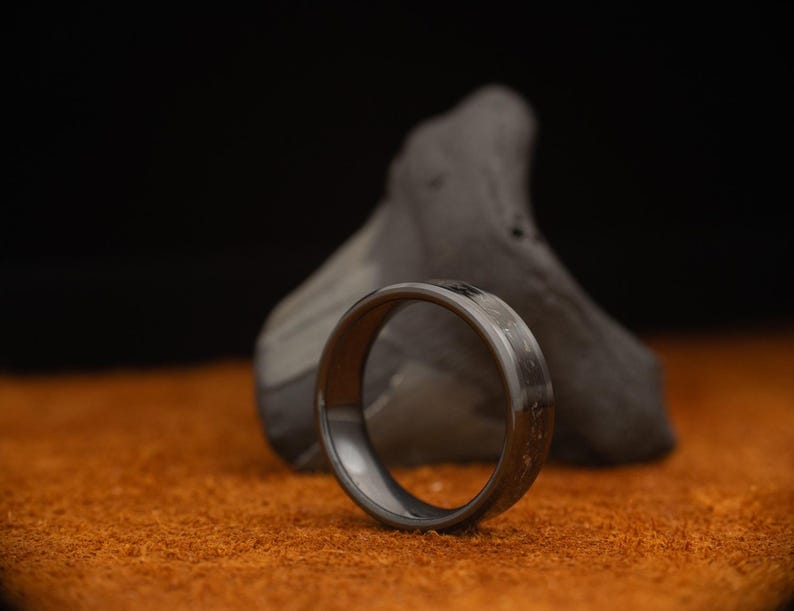 Peut inclure: Une bague grise fonc&eacute;e avec une surface textur&eacute;e, sur un fond flou. La bague est faite d'un mat&eacute;riau sombre et repose sur une surface orange. La bague est un bijou.