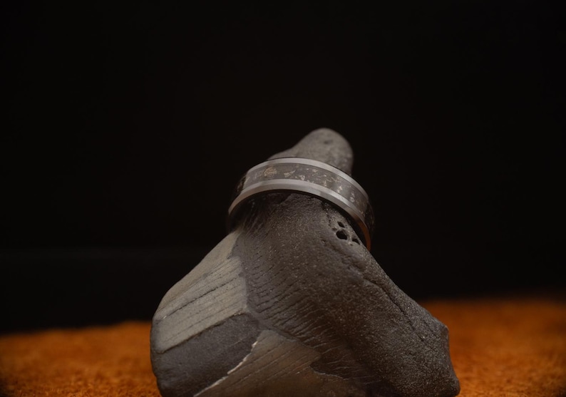 Peut inclure: Gros plan d'une bague argent&eacute;e avec une bande sombre et textur&eacute;e. La bague est mise en valeur sur un fond sombre, soulignant son &eacute;clat m&eacute;tallique et son design complexe. La bague repose sur une pierre grise fonc&eacute;e.