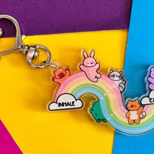 Puede incluir: Un llavero de acrílico con un arcoíris y un cierre en forma de corazón. El diseño incluye un conejo rosa, un oso marrón, un mapache, una rana verde y un gato naranja. Las nubes blancas tienen impreso el texto INHALE a la izquierda y EXHALE a la derecha.