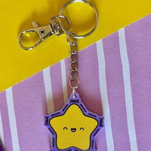 May include: A yellow star-shaped keyring with a smiling face and the words "HOLD" and "BREATHE OUT" in purple. The keyring has a silver clasp and chain, set against a yellow and purple background.