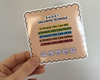 May include: A pink and white sticker with a grounding technique chart. The chart lists 5 things you can see, 4 things you can touch, 3 things you can hear, 2 things you can smell, and 1 thing you can taste. The sticker has a scalloped border and four cartoon pig faces at the bottom.
