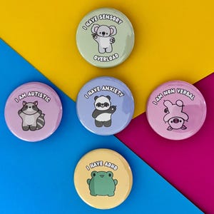Peut inclure: Plusieurs badges ronds et colorés avec des illustrations d'animaux de dessins animés. Chaque badge porte une phrase différente, notamment "I AM AUTISTIC", "I HAVE SENSORY OVERLOAD", "I HAVE ANXIETY", "I AM NON VERBAL" et "I HAVE ADHD".