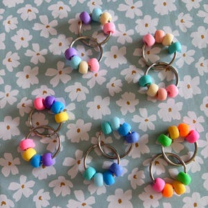 May include: A selection of beaded keyrings in various pastel colours. Each keyring has a silver ring with a series of beads in a circular pattern. The beads are in shades of pink, yellow, blue, and purple.