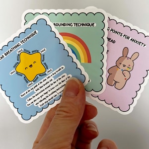 Peut inclure: Trois cartes carrées aux bords festonnés sont tenues dans une main. Les cartes ont des motifs et du texte différents. Une carte présente une étoile jaune avec le texte "STAR BREATHING TECHNIQUE". Une autre carte présente un arc-en-ciel et le texte "ROUNDING TECHNIQUE". La troisième carte présente un lapin et le texte "G POINTS FOR ANXIETY".
