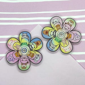 Puede incluir: Dos fidget spinners con forma de flor y fondo degradado en amarillo, azul y morado. Cada pétalo muestra un animal de dibujos animados diferente, incluyendo un mapache, panda, koala, rana, cerdo y ajolote, con un rodamiento circular central.