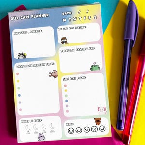 Op de afbeelding: Een zelfzorgplanner met een pastelkleurige gradiëntachtergrond, met secties voor dagelijkse affirmaties, dankbaarheid en het volgen van de stemming. Bevat schattige dierenillustraties en de tekst "SELF CARE PLANNER". Twee pennen, paars en rood, zijn rechts geplaatst.