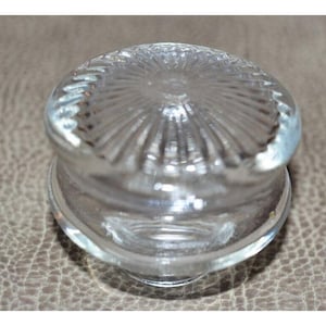 May include: A clear glass bottle stopper with a circular, ridged design on the top. The stopper has a rounded shape and is made of transparent glass. The design is reminiscent of a sunburst.