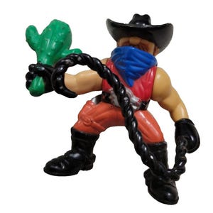 May include: A plastic action figure of a cowboy wearing a black hat, a blue bandana, a red sleeveless top, and orange pants. The figure has black boots and gloves, holds a green cactus in one hand, and wields a black coiled whip in the other.