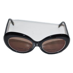 May include: Black oval-shaped sunglasses with brown lenses. The sunglasses have a thick, glossy black frame and a classic, retro design. The lenses are a warm brown color, offering a contrast to the black frame.