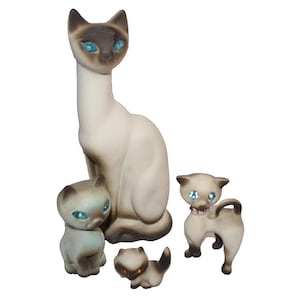 May include: A set of four Siamese cat figurines with cream-colored bodies and dark brown points. The cats feature bright blue or amber rhinestone eyes. One cat wears a small collar with a pink charm. The figures vary in size and pose.