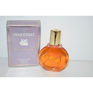 May include: A Vanderbilt eau de toilette spray bottle with orange liquid and a gold cap, featuring a molded glass swan design. Beside it stands a rectangular lavender box with gold trim, displaying the text Vanderbilt and a swan graphic.