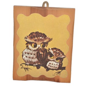 May include: A rectangular wooden wall plaque featuring a painted illustration of two brown and white owls perched on a branch. The owls have large yellow eyes and are set against a scalloped, mustard-yellow background. A small leather loop is attached to the top.