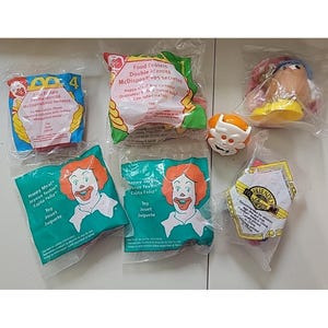 May include: A collection of sealed McDonald's Happy Meal toys, including a Mr. Potato Head, a white-faced character, and several packages featuring Ronald McDonald. The packages have text in multiple languages.