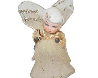 Pode incluir: Uma estatueta de anjo em cerâmica com cabelo branco, olhos azuis e um vestido cor de creme feito de tecido de tule em camadas. O anjo apresenta asas grandes e transparentes com detalhes de glitter e segura uma pequena varinha castanha em estrela.