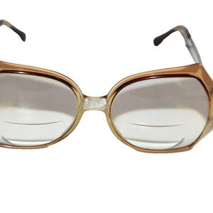 May include: Vintage eyeglasses with a light brown, angular frame and clear lenses. The glasses have a bifocal design, with a subtle line visible across the lower portion of each lens. The temples are a light brown and black pattern.