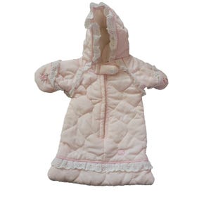 Peut inclure: Une gigoteuse matelassée rose pâle dotée d'une capuche avec une bordure en dentelle blanche à œillets. Le vêtement comprend des manches longues avec de petites broderies florales, une fermeture éclair frontale et un volant en dentelle.