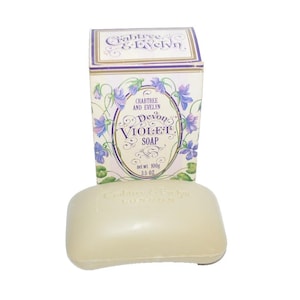 May include: A bar of Devon Violet Soap from Crabtree & Evelyn, next to its matching box. The soap is a light beige color, and the box features a floral design with the brand name and product details. The box states the net weight is 3.5 oz.