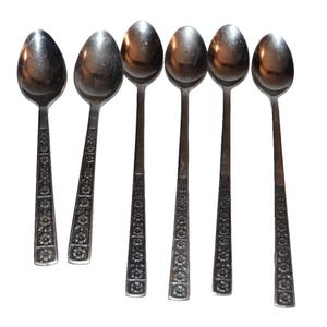 Könnte beinhalten: Ein Set aus sechs silberfarbenen Metalllöffeln, die in einer Reihe angeordnet sind. Jeder Löffel hat einen langen, schlanken Griff, der mit einem sich wiederholenden geprägten Blumenmuster verziert ist, das vom Laffe bis zum Griffende reicht.