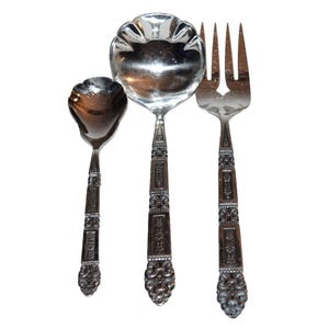 May include: Three pieces of ornate silverware, including a serving spoon, a smaller spoon, and a fork. The silverware is silver-coloured with detailed decorative handles. The serving spoon has a scalloped edge.
