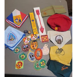 Peut inclure: Une collection de souvenirs scouts vintage comprenant divers écussons brodés, des foulards, un béret rouge avec un emblème fleur de lys, une casquette de garnison beige et une longue écharpe blanche avec un motif de flèche rouge et des insignes.