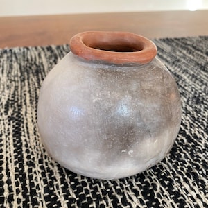 Puede incluir: Un pequeño jarrón redondo de cerámica gris con un borde marrón. El jarrón está sobre una alfombra tejida en blanco y negro.