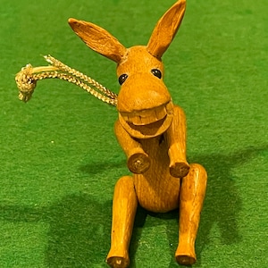 May include: A wooden donkey ornament with a wide grin and black eyes. The donkey is sitting with its legs outstretched and has a gold cord attached to its back.