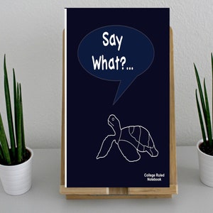 May include: A navy blue college ruled notebook featuring a white turtle illustration and the text "Say What?..." in a speech bubble. The notebook is displayed on a wooden stand, with two potted snake plants on either side.