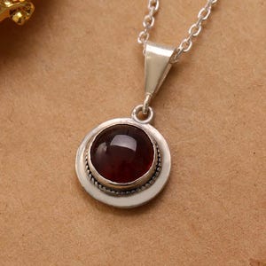 May include: A silver necklace with a round, dark red gemstone pendant. The pendant is set in a silver bezel with a beaded edge. The chain is a delicate silver link. The necklace is displayed on a brown surface.