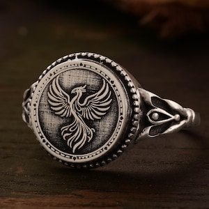 May include: A silver ring with a round, detailed design featuring a phoenix bird. The ring has a textured, ornate band.