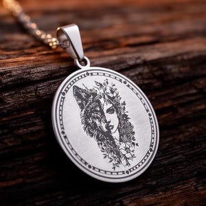 Puede incluir: Collar con colgante de plata con diseño grabado. El colgante redondo muestra una imagen mitad lobo, mitad mujer. La cadena es de eslabones finos de plata. El fondo es una superficie de madera oscura y texturizada.