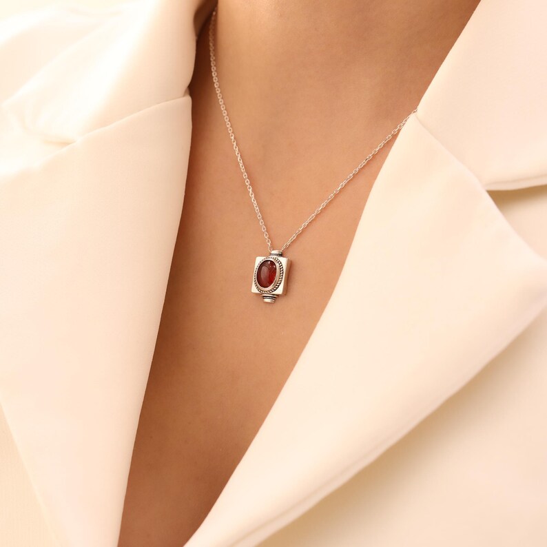 May include: A silver necklace with a rectangular pendant featuring a red gemstone. The pendant is framed by a decorative border and hangs from a delicate chain. The necklace is displayed against a cream-colored background.