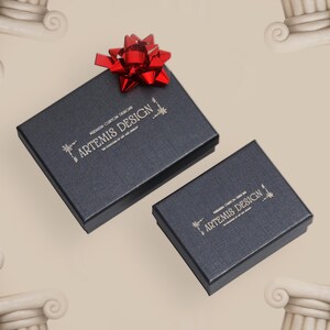 May include: Two dark gray gift boxes with the text "ARTEMIS DESIGN" in gold lettering. A bright red bow sits atop the larger box. The boxes are on a light beige surface.