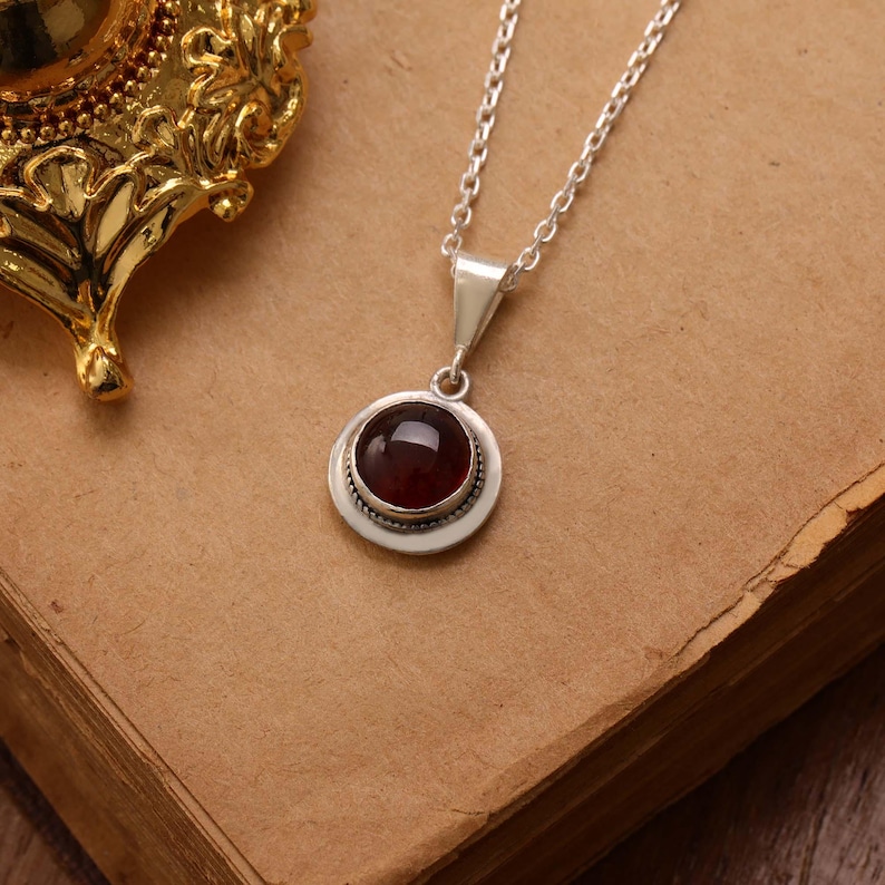 May include: A silver necklace with a round, dark red gemstone pendant. The pendant is set in a silver bezel with a beaded edge. The chain is a delicate silver link. The necklace is displayed on a brown surface.