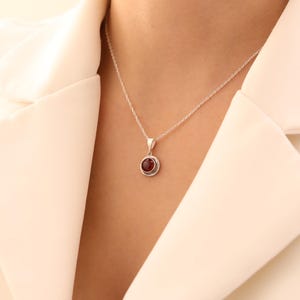 May include: A silver necklace with a small, round, red gemstone pendant. The pendant is set in a detailed silver bezel. The necklace is displayed against a white background.