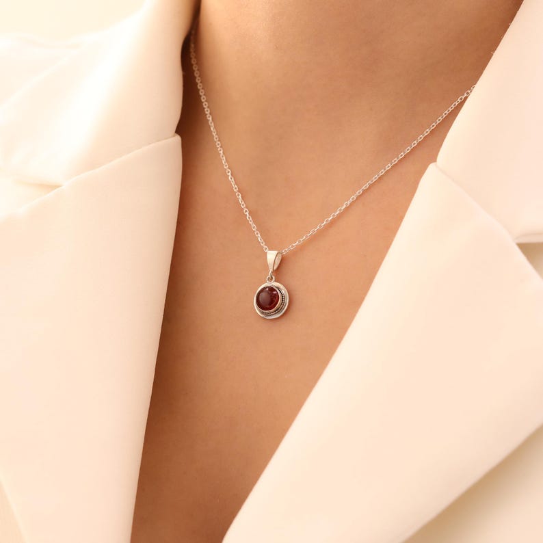 May include: A silver necklace with a small, round, red gemstone pendant. The pendant is set in a detailed silver bezel. The necklace is displayed against a white background.