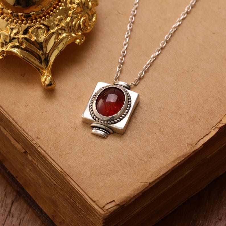 May include: A silver necklace with a square pendant featuring a red gemstone. The gemstone is oval-shaped and surrounded by a twisted rope design. The pendant hangs from a delicate silver chain, resting on a vintage book.