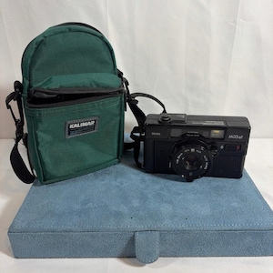 May include: A black Sears M35af 35mm film camera with a lens featuring Auto Sears text, positioned next to a green Kalimar camera bag with a black strap. The camera has a rectangular body with a top-mounted shutter button and a front-facing lens.